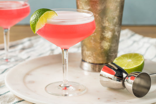 A pink Cosmopolitan cocktail on a white table, with a lime wedge garnish. It's next to a cocktail shaker and a jigger.