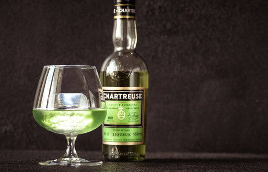 Green Chartreuse bottle next to a glass of Chartreuse green liqueur with an ice cube in it, on a black background