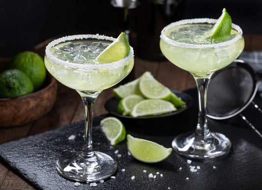 Two margarita cocktails sitting on a black chopping board. The cocktails have a salt rim and lime wedges. The chopping board has more lime wedges on it.